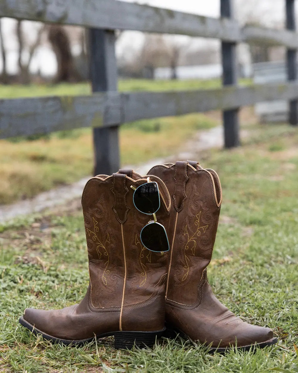 A pair of brown cowboy boots sits on grass near a wooden fence. A pair of reflective sunglasses hangs from one boot. The background shows blurred trees and more fence on a cloudy day.