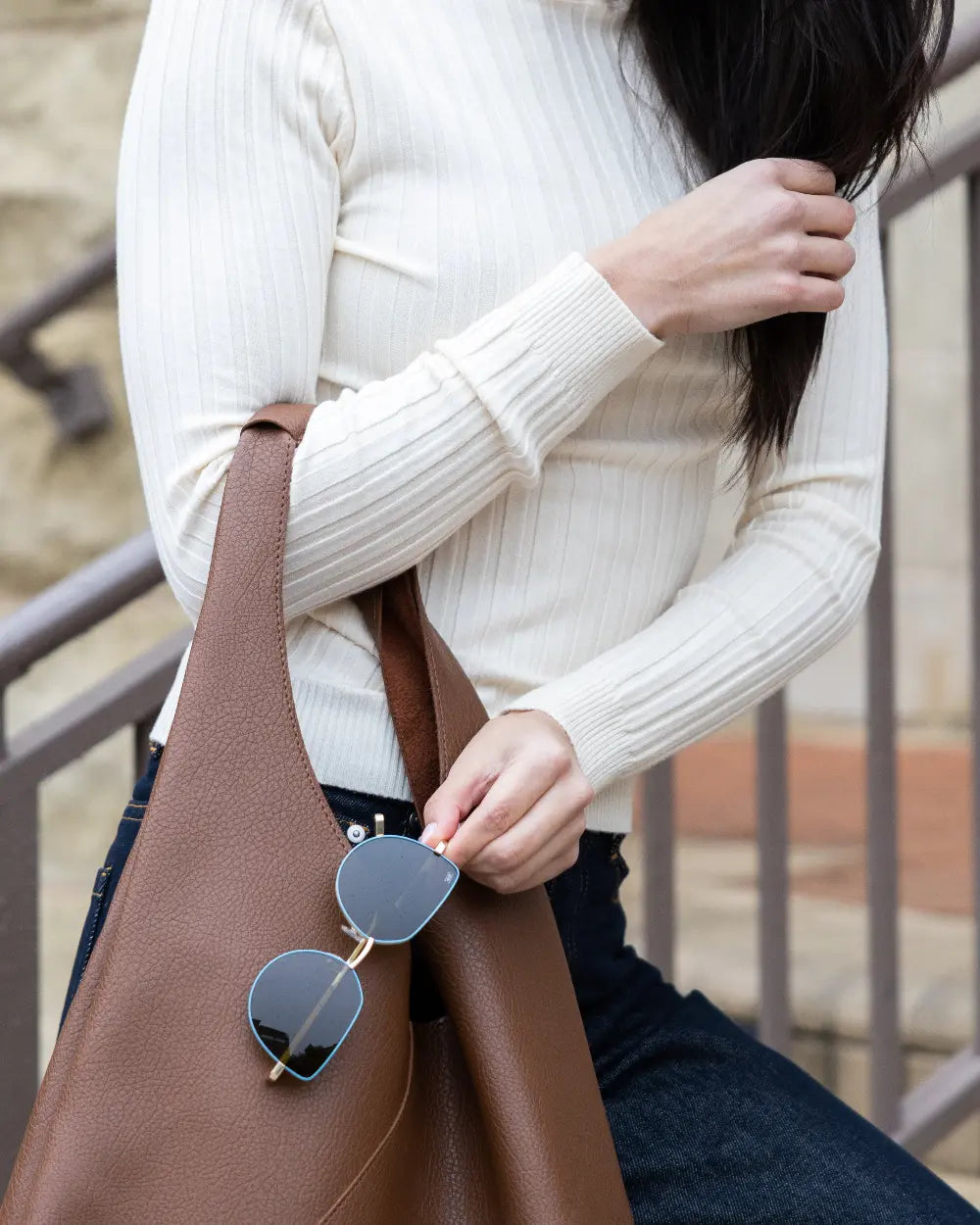 A person wearing a cream ribbed turtleneck sweater and jeans holds a large brown leather bag. They are holding sunglasses and standing near a railing, with their long dark hair partially covering their face.