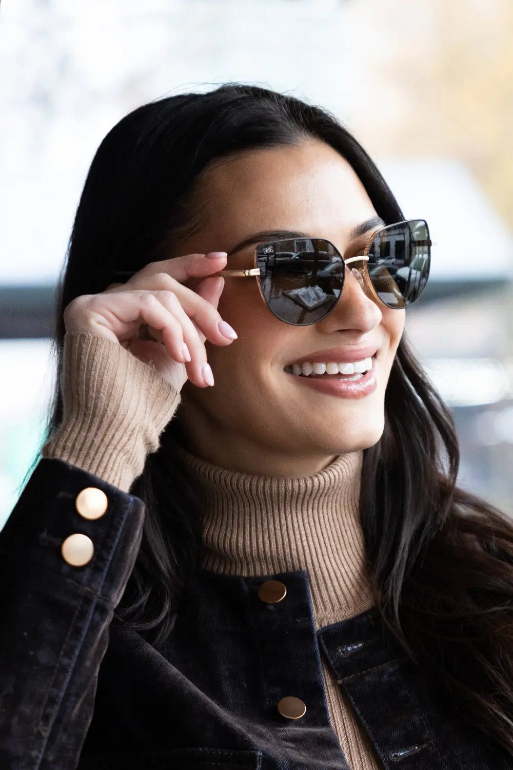 A person with long dark hair smiles while wearing sunglasses and a beige turtleneck sweater. The person holds the sunglasses with one hand. They are outdoors with a blurred background.