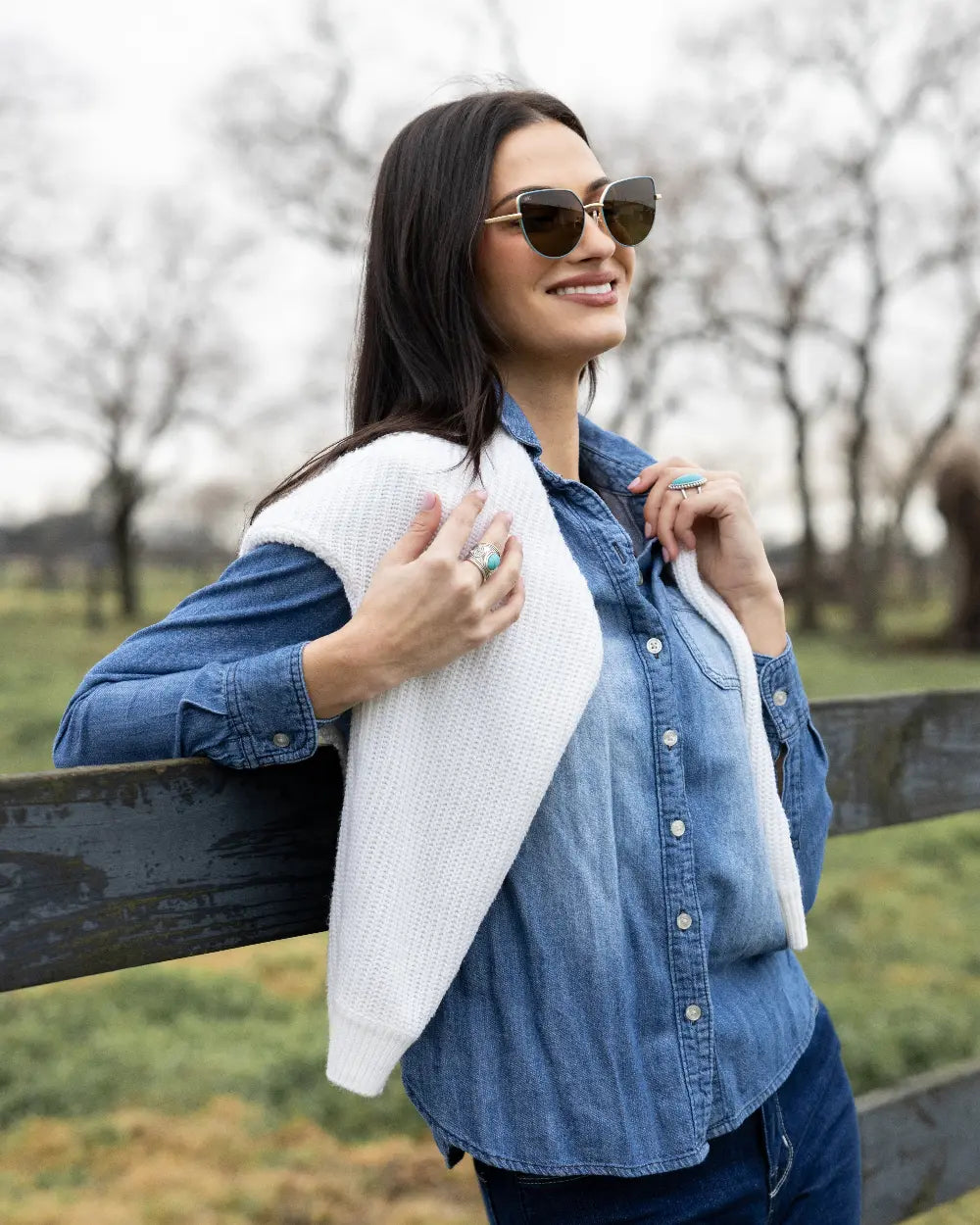 Woman in sunglasses and denim shirt leans on a wooden fence, smiling. A white sweater is draped over her shoulders. Shes outdoors in front of a blurred background of trees and grass on a cloudy day.