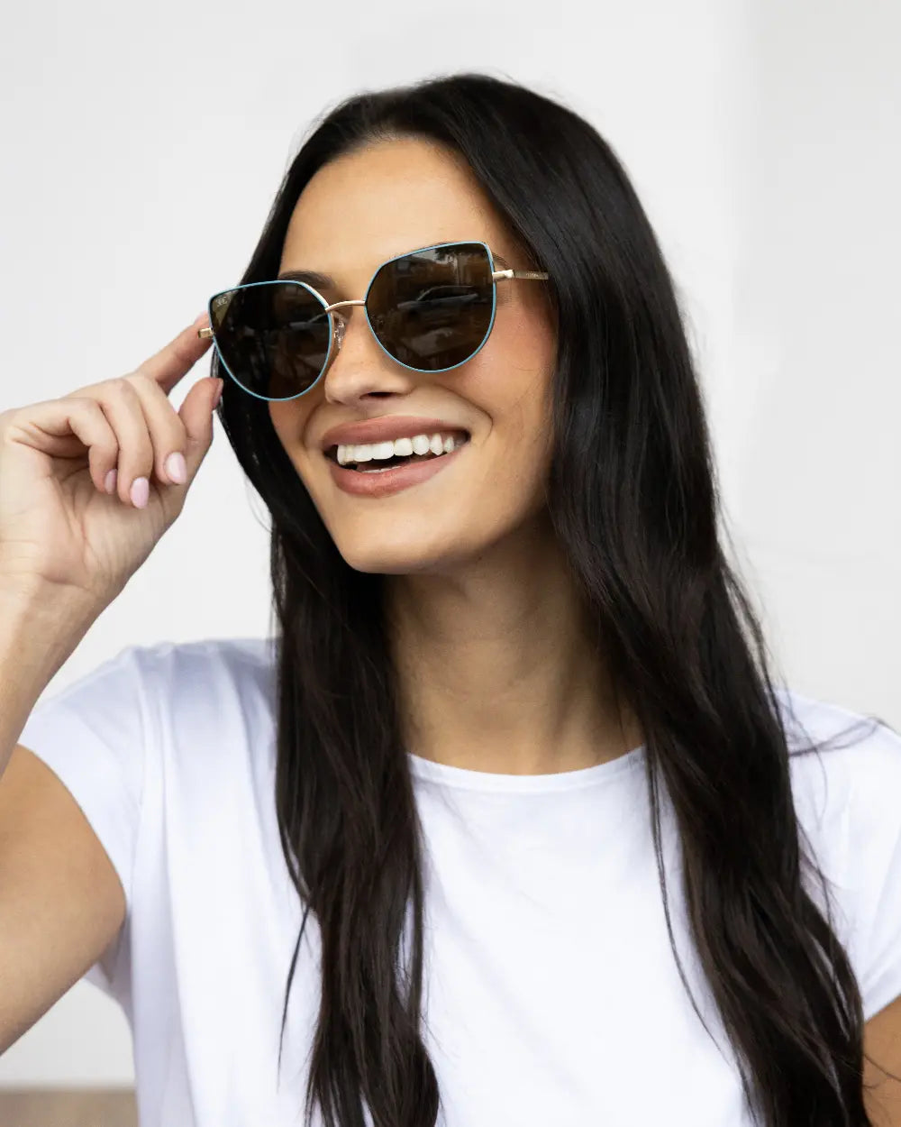A woman with long dark hair smiles while adjusting her sunglasses. She is wearing a white t-shirt and is standing against a plain white background.
