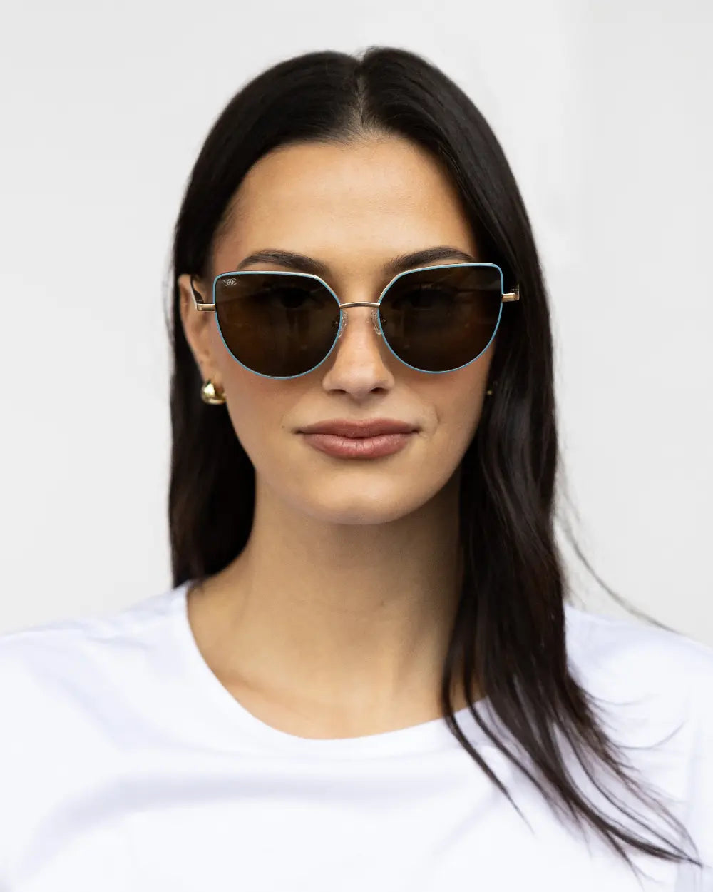 Person with long dark hair wearing sunglasses and a white shirt, facing forward. They have a neutral expression and are standing against a plain light-colored background.