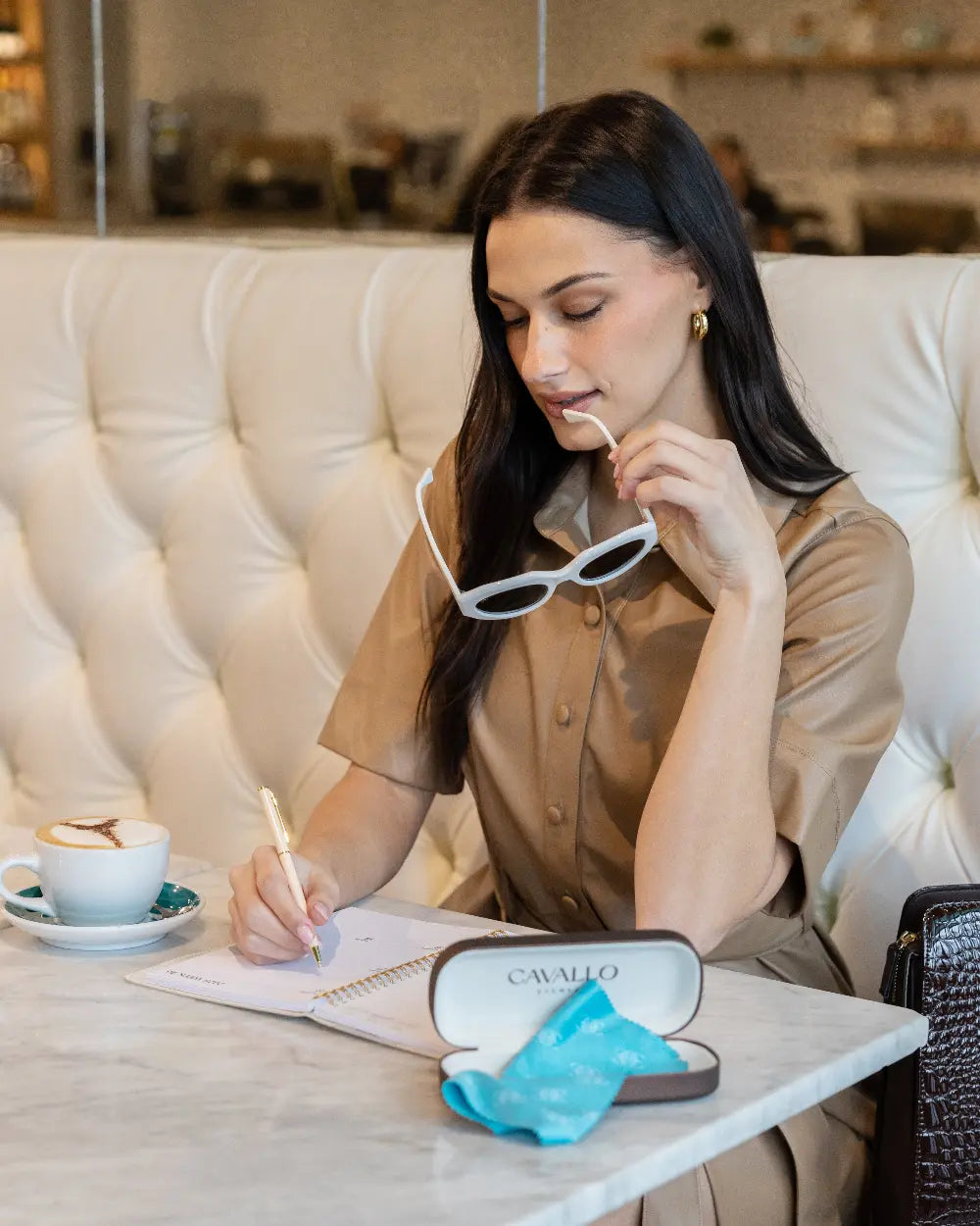 A woman with long dark hair sits at a cafe table, holding white sunglasses and writing in a notebook. A cup of coffee and a case labeled Cavallo with a blue cloth are on the table. She wears a beige dress and gold hoop earrings.