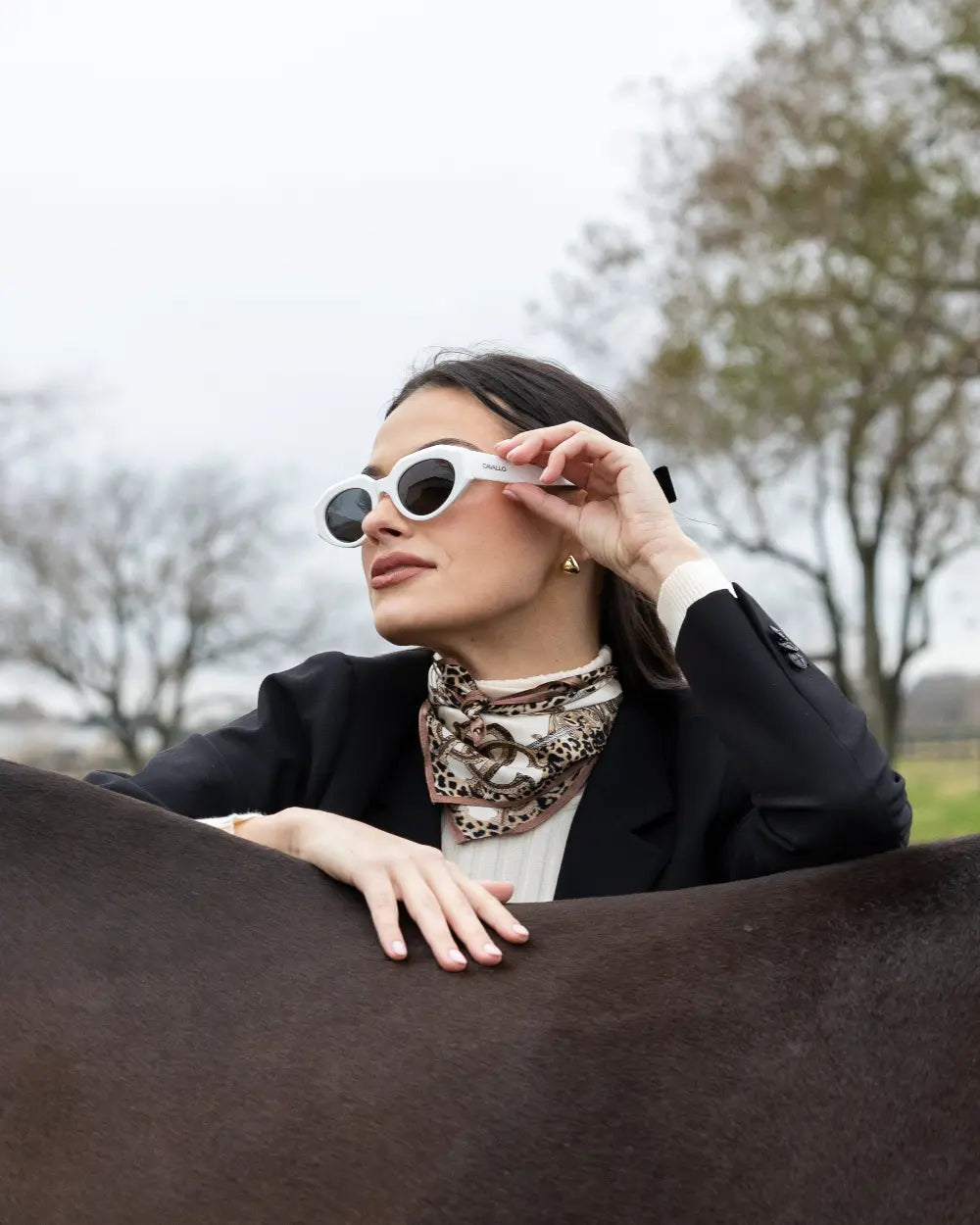 A woman in a black blazer and patterned scarf stands beside a brown horse, holding white-framed sunglasses with one hand. She is outdoors with bare trees in the background, looking off into the distance.