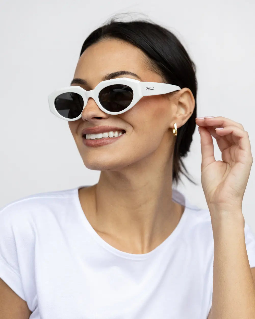 Smiling person wearing white sunglasses and a white t-shirt. They have dark hair pulled back and are touching their ear, showcasing a small hoop earring. The background is plain and light-colored.