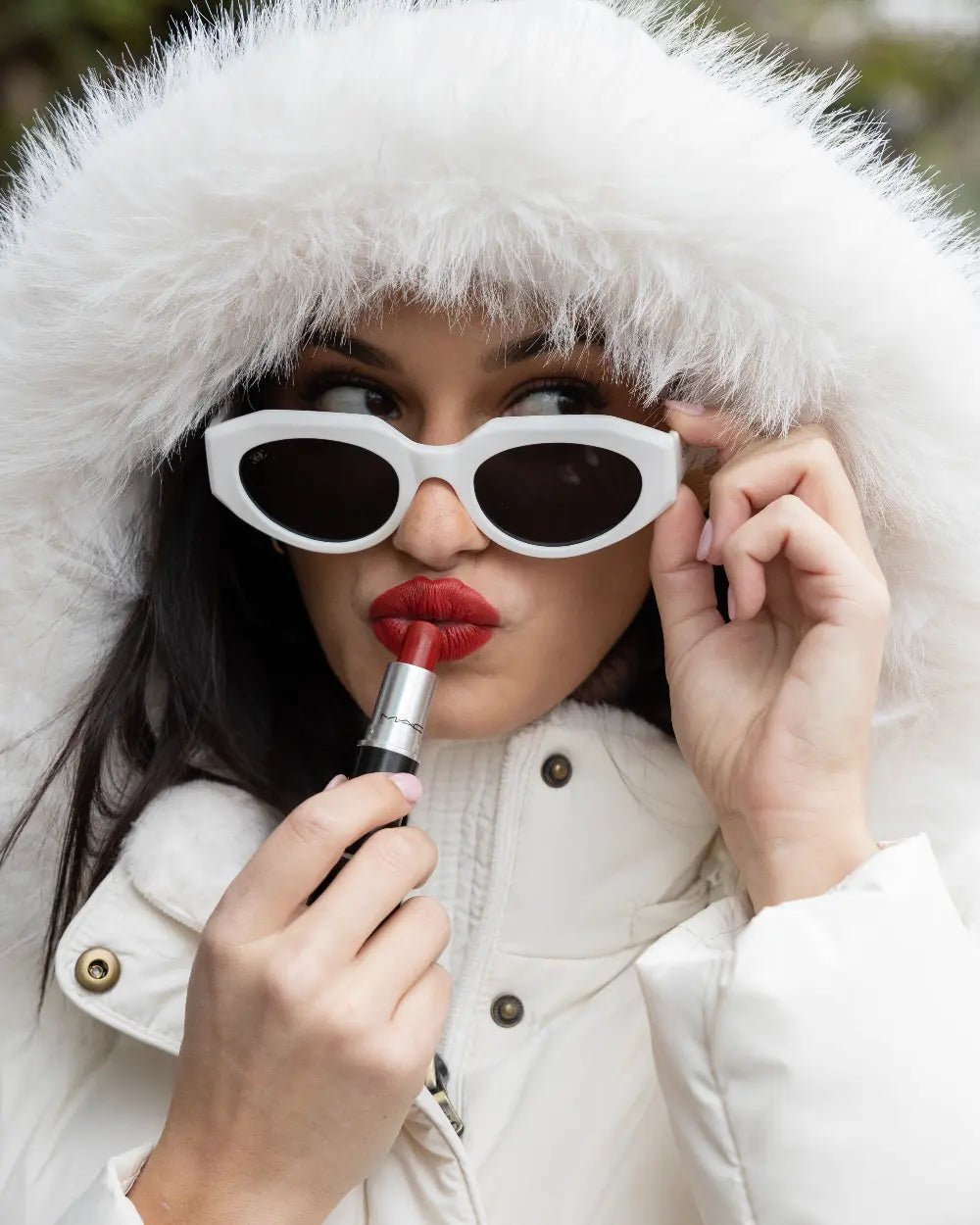 A person wearing a white fur-lined hooded coat and white cat-eye sunglasses applies red lipstick. They hold the sunglasses with one hand and look thoughtfully to the side. The background is blurred greenery.