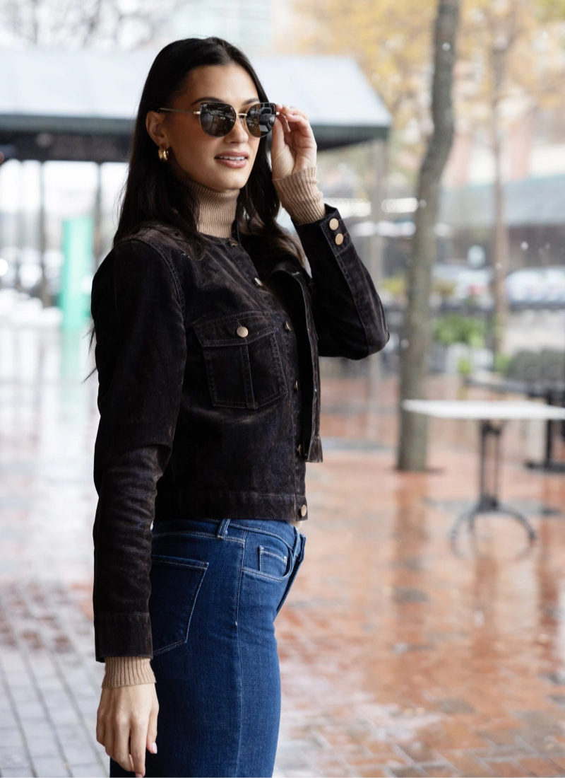 Woman in sunglasses and a dark jacket poses confidently outdoors on a rainy day. She stands on a wet brick pavement with blurred trees and buildings in the background.