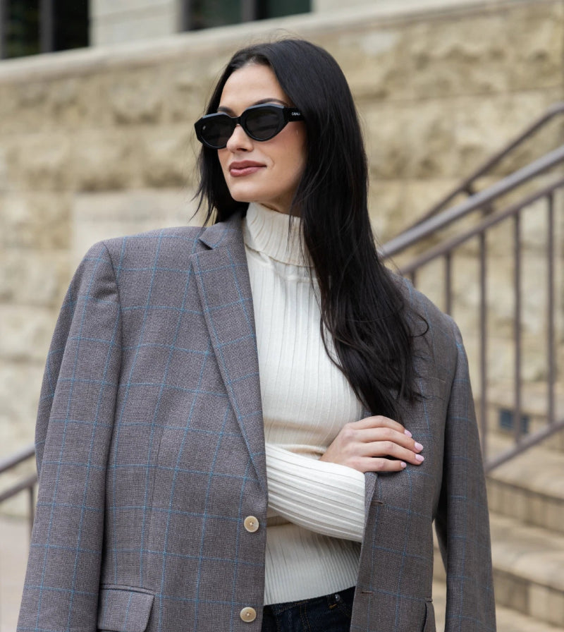 A woman wearing a large gray plaid blazer over a white turtleneck stands confidently. She has long dark hair and is wearing sunglasses. The background features stone steps and a wall.