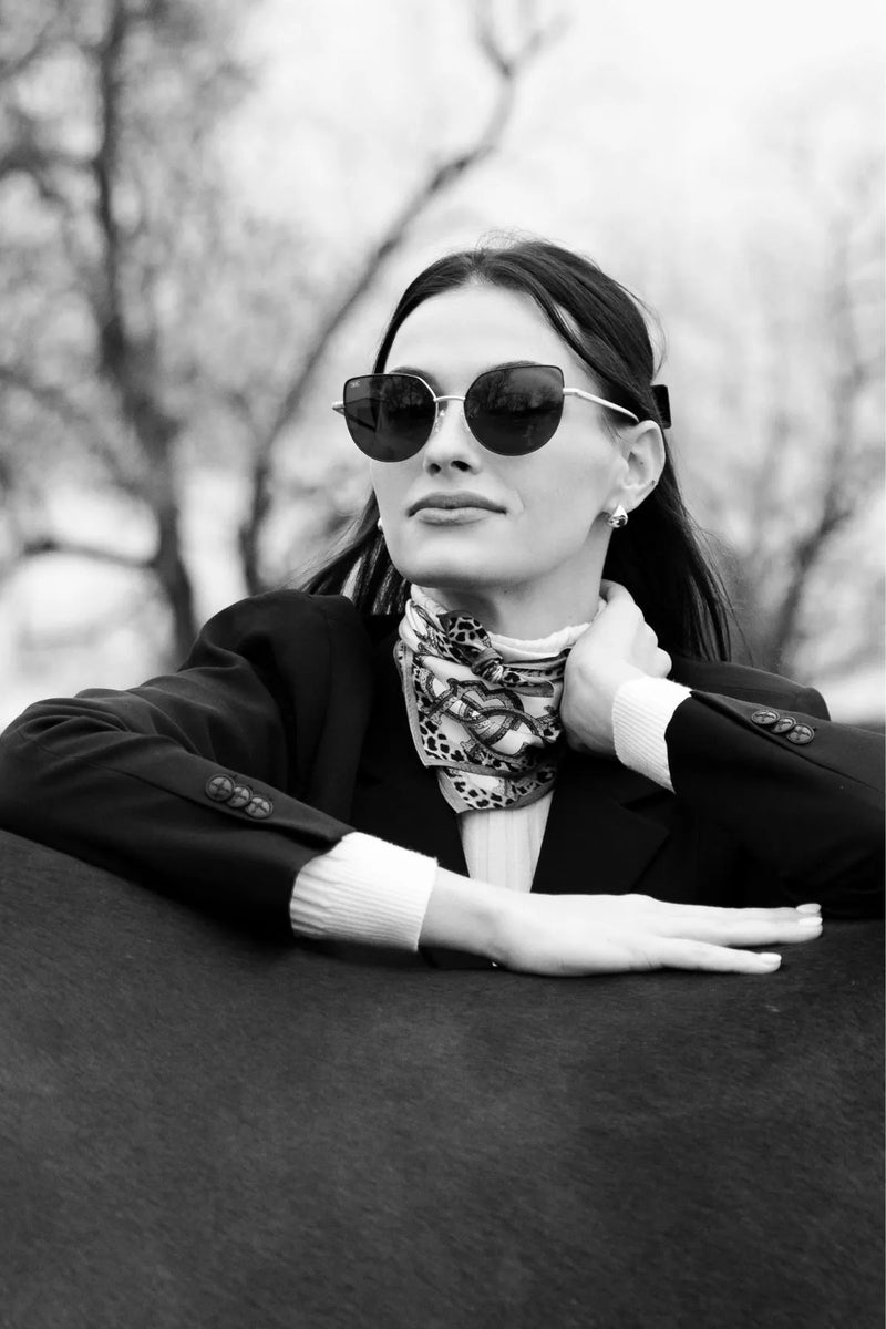Elegant woman in sunglasses and a scarf leaning on a horse, exuding luxury and sophistication in an outdoor setting.